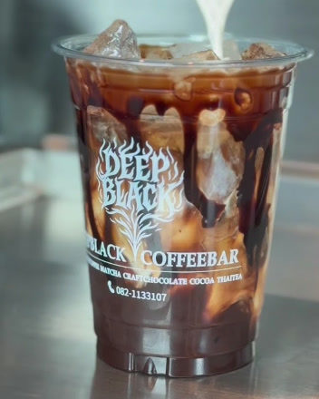 เทนมลงในแก้วกาแฟเย็นที่มีโลโก้ DEEP BLACK บนเคาน์เตอร์สเตนเลส